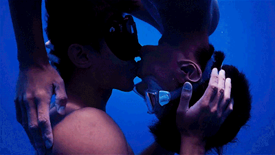 Two people kissing underwater, both wearing diving masks.