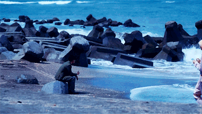 Person sitting on a rock at a rocky beach, looking down toward the shore as blue waves roll in.