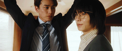 Two coworkers in suits in an office; the man on the left raises his arms while the man on the right watches.