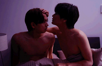 Two topless people in bed, facing each other as one gently cups the other's head, leaning in for a kiss-like moment.