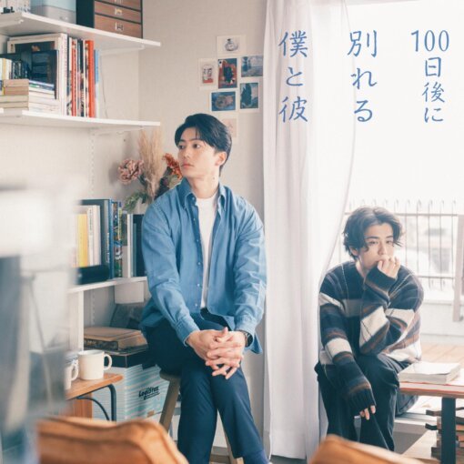 Two men in a bright study: one in a blue shirt sits on a stool, the other in a striped sweater near a window with a curtain featuring blue Japanese text plus shelves of books and boxes in the background.