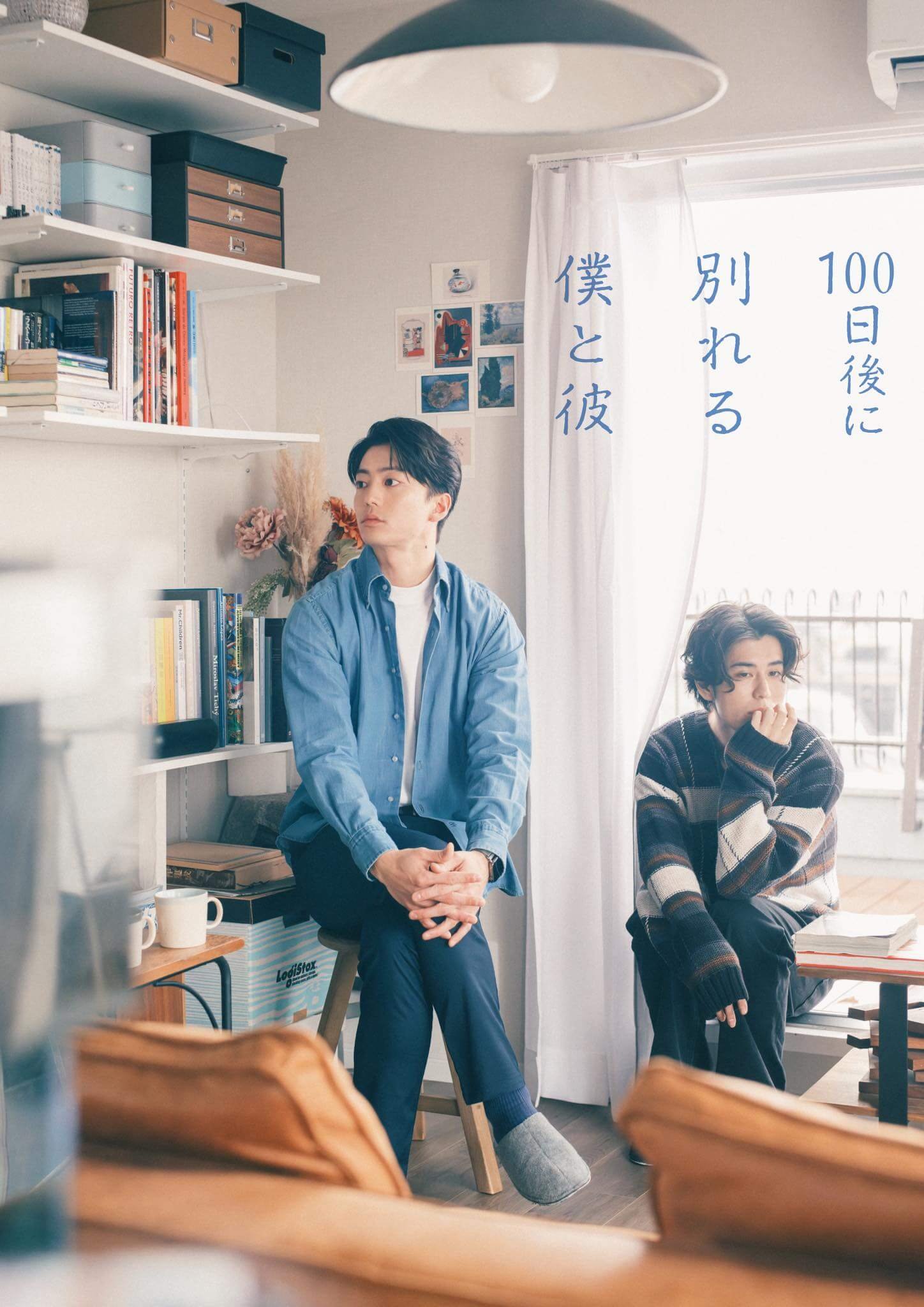 Two men in a bright study: one in a blue shirt sits on a stool, the other in a striped sweater near a window with a curtain featuring blue Japanese text plus shelves of books and boxes in the background.