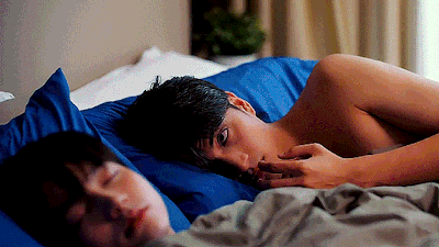 Man lying shirtless in bed, awake and looking toward the camera while a woman sleeps beside him in a dimly lit room.
