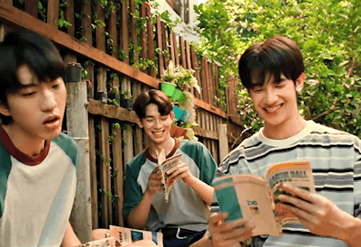Three young friends outdoors by a wooden fence, reading books and sharing a smile.