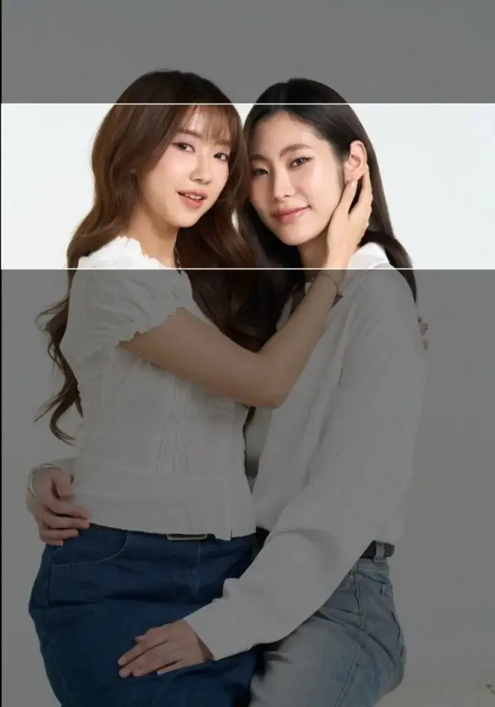 Two women posing closely together, smiling at the camera with their arms around each other, wearing white tops and jeans (friendly portrait).