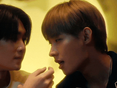 Two young men in a yellow-lit room, facing each other as they talk.