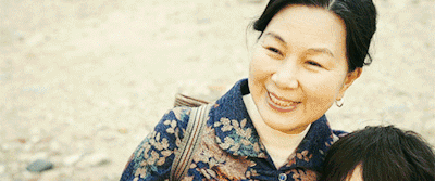 Smiling Asian woman in a blue floral-pattern shirt on a beach, with a second person partially visible beside her.