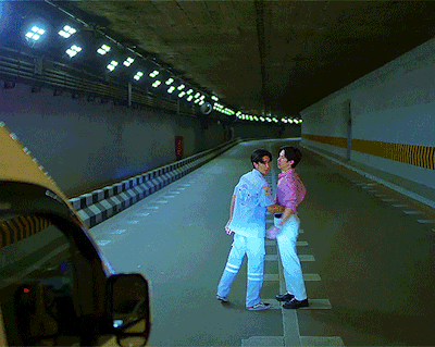 Two men face each other in a dim tunnel; one wears a light blue suit, the other a pink shirt, with a car’s side mirror visible in the foreground.