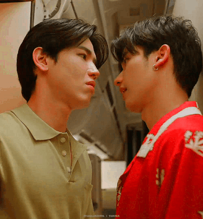 Two young men stand inches apart, facing each other with intense expressions in a hallway, one in a beige polo and the other in a red jacket.