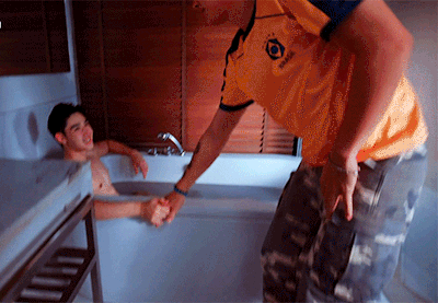 A man in a yellow shirt reaches over a bathtub edge to grab and pull up another man who is sitting inside the tub.