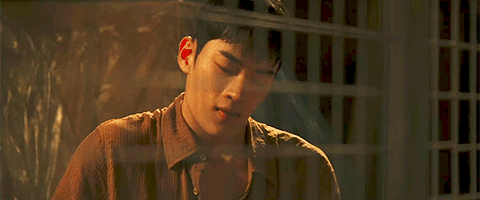Young man in a brown shirt looking down, seen through a glass window with reflections.
