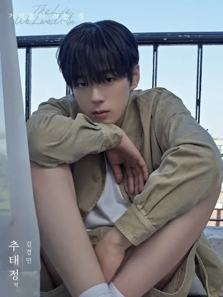 Portrait of a young man with dark hair sitting with knees up, wearing a beige jacket and white shirt, gazing at the camera outdoors.