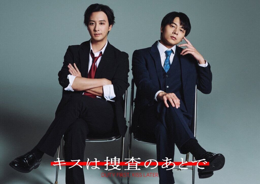 Two men in suits sit on chairs against a gray backdrop, legs crossed, posing confidently; poster text at bottom reads 'Duty First, Kiss Later' (in red).