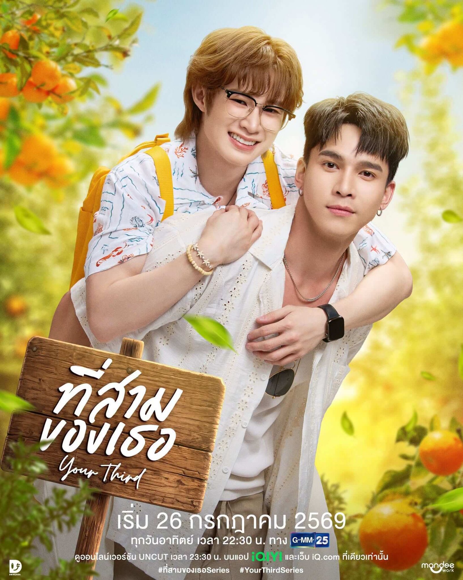 Two young men outdoors, one giving the other a piggyback, smiling, holding a wooden sign with Thai script amid bright orange trees and greenery.